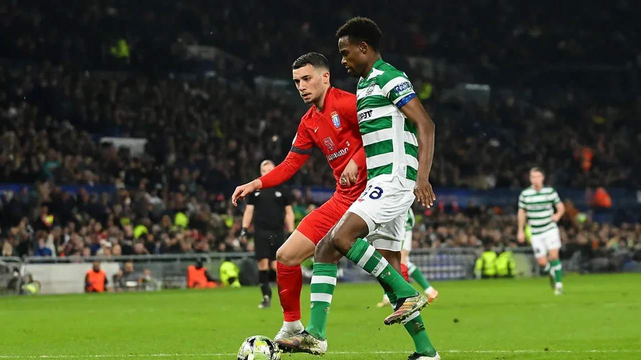 Dois jogadores em duelo ombro a ombro pela bola, um de vermelho e outro de verde e branco, com adeptos nas bancadas