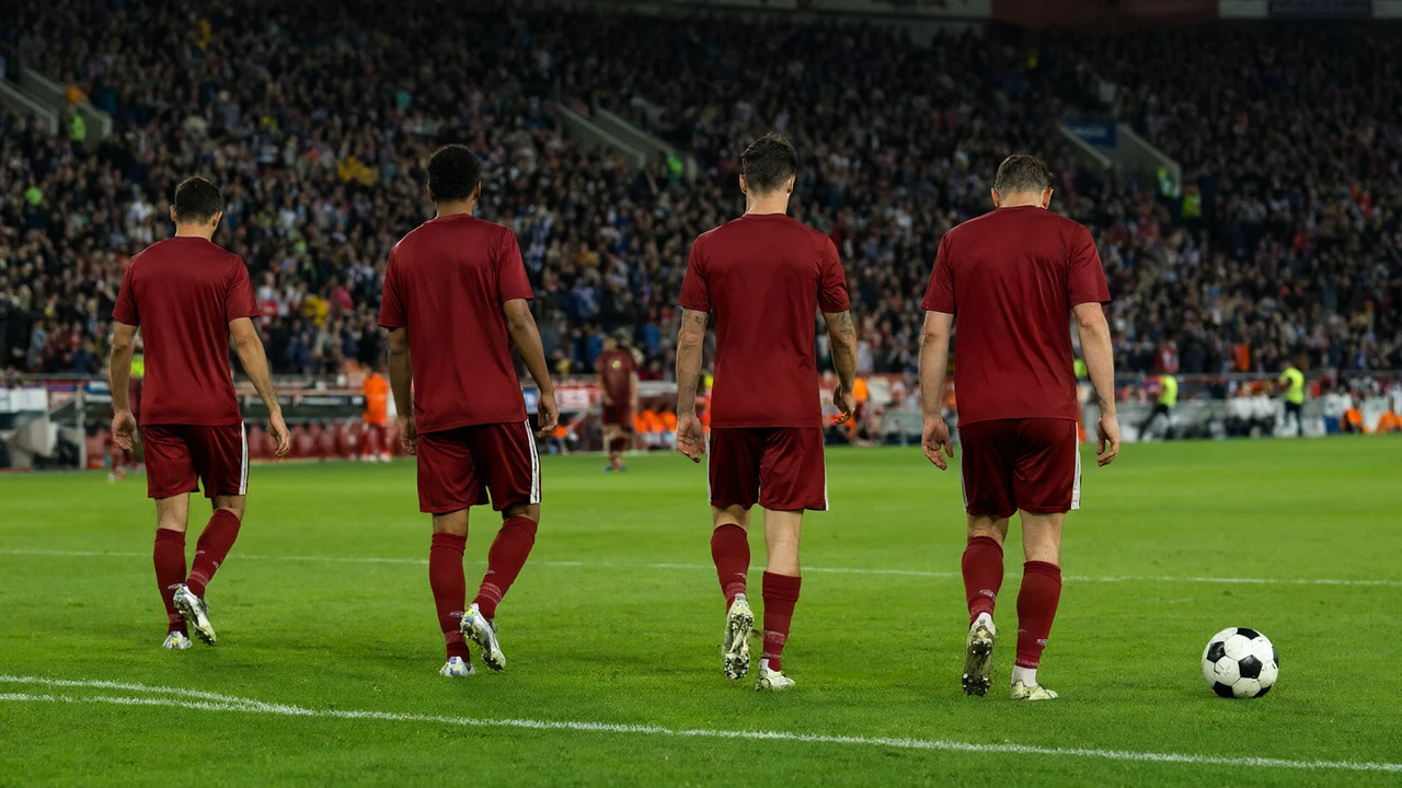 Jogadores de vermelho a sair do relvado cabisbaixos depois de uma derrota, com o estádio iluminado ao fundo
